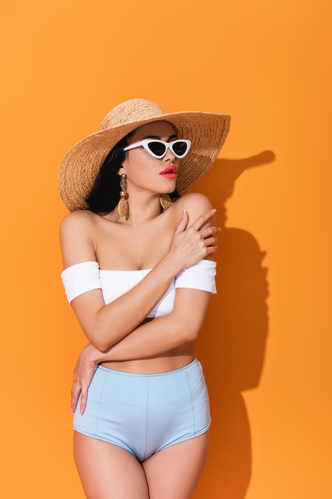 стильная женщина в купальнике, солнцезащитных очках и соломенной шляпе, стоящая со скрещенными руками на оранжевом
  - Фото, изображение