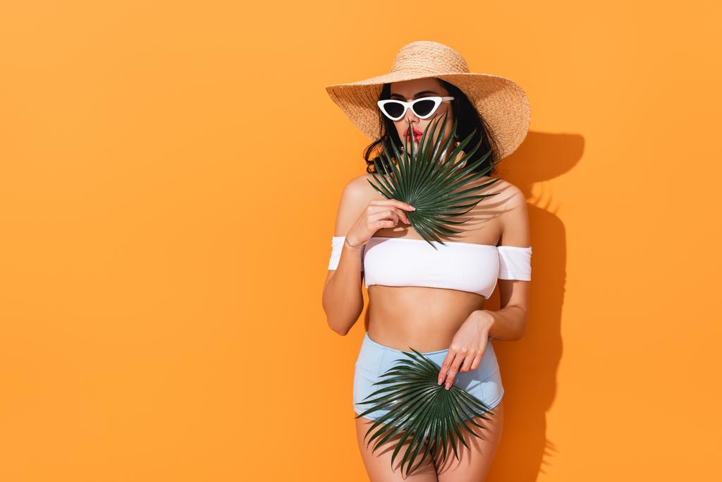 стильная женщина в купальнике, солнцезащитных очках и соломенной шляпе держит пальмовые листья на оранжевом
 - Фото, изображение