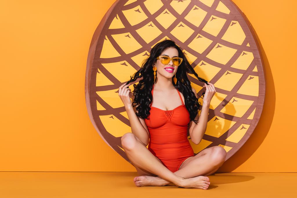 attraente e scalza donna in costume da bagno e occhiali da sole seduto in posa yoga vicino ananas taglio di carta su arancione, concetto estivo  - Foto, immagini