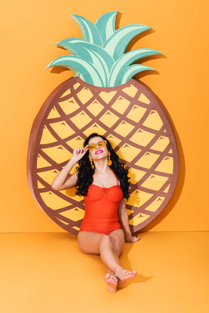happy and barefoot woman in swimsuit sitting near paper cut pineapple and touching sunglasses on orange, summer concept  - Photo, Image