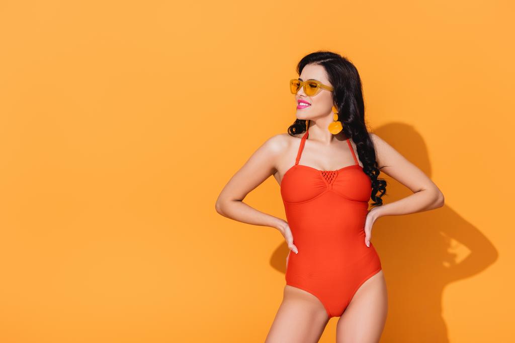 szczęśliwa kobieta w strojach kąpielowych i okularach przeciwsłonecznych stojąca z dłońmi na biodrach na pomarańczowym  - Zdjęcie, obraz