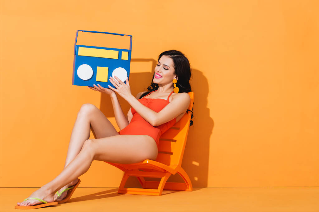 cheerful young woman in swimsuit holding paper boombox while sitting on deck chair on orange - Photo, Image