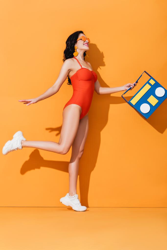 Χαρούμενη γυναίκα με αθλητικά παπούτσια, γυαλιά ηλίου και μαγιό που κρατάει χάρτινο κουτί σε πορτοκαλί - Φωτογραφία, εικόνα