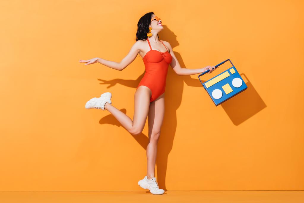 щаслива жінка в кросівках, сонцезахисних окулярах та купальнику, що тримає паперову коробку на апельсині
 - Фото, зображення