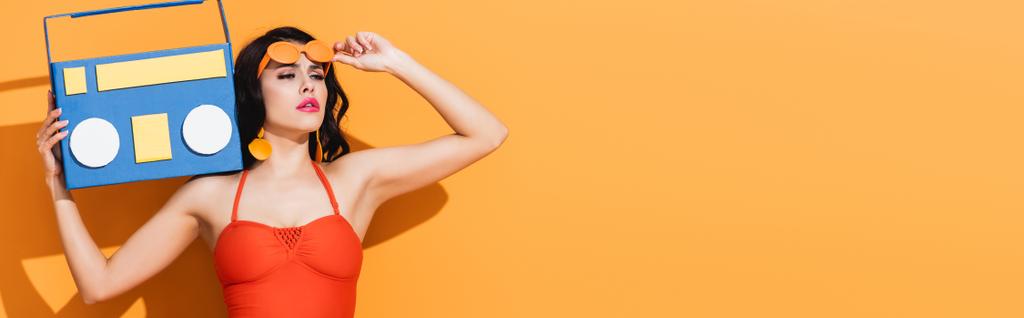 raccolto panoramico di donna elegante in costume da bagno toccare occhiali da sole mentre tiene carta tagliata boombox su arancione - Foto, immagini