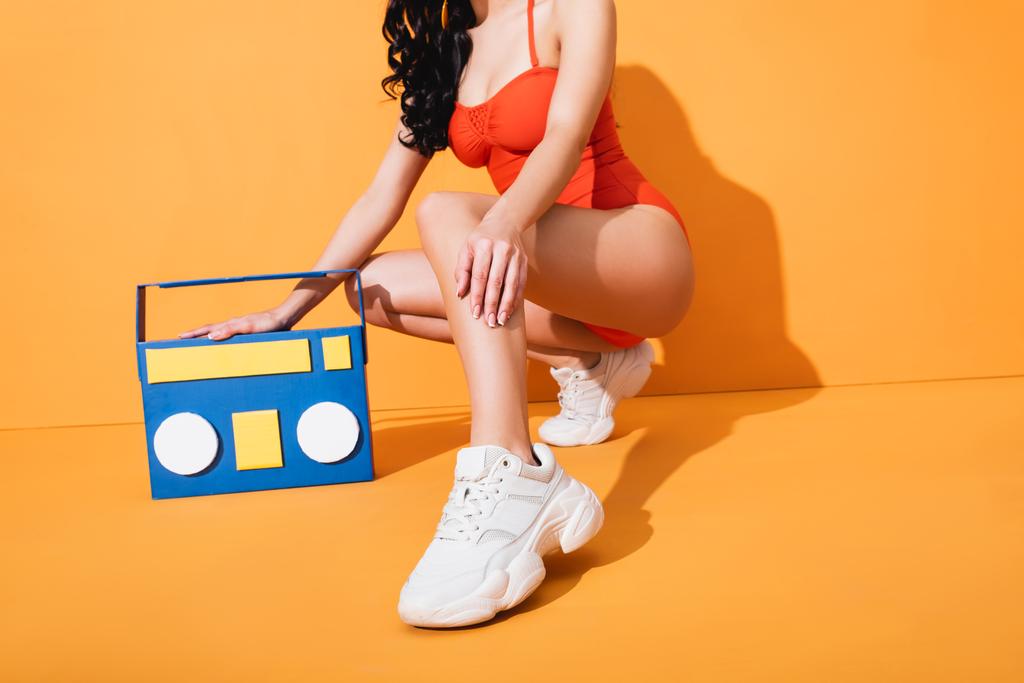 обрізаний вид стильної жінки в кросівках і купальнику, що сидить біля паперової коробки на помаранчевому
 - Фото, зображення