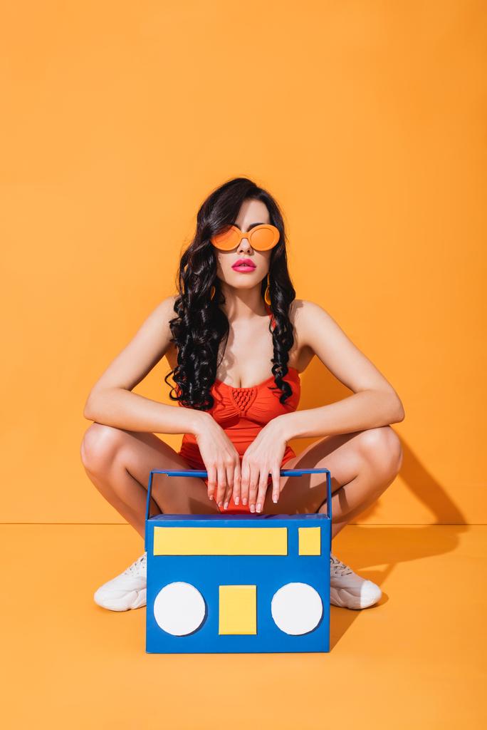 stijlvol meisje in sneakers, badpak en zonnebril zit in de buurt van papier gesneden boombox op oranje - Foto, afbeelding