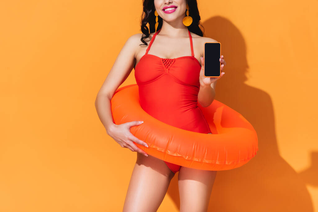przycięty widok szczęśliwej kobiety w stroju kąpielowym z nadmuchiwanym pierścieniem trzymając smartfon z pustym ekranem na pomarańczowym - Zdjęcie, obraz