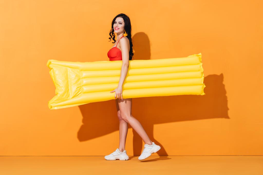 ragazza felice in costume da bagno e scarpe da ginnastica che tengono il materasso gonfiabile su arancione  - Foto, immagini