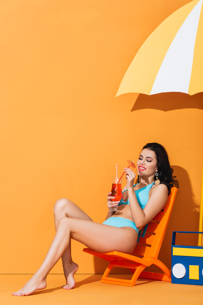 uśmiechnięta kobieta w stroju kąpielowym siedząca na leżaku przy papierowej boomboxie i parasolce patrząc na koktajl na pomarańczy - Zdjęcie, obraz