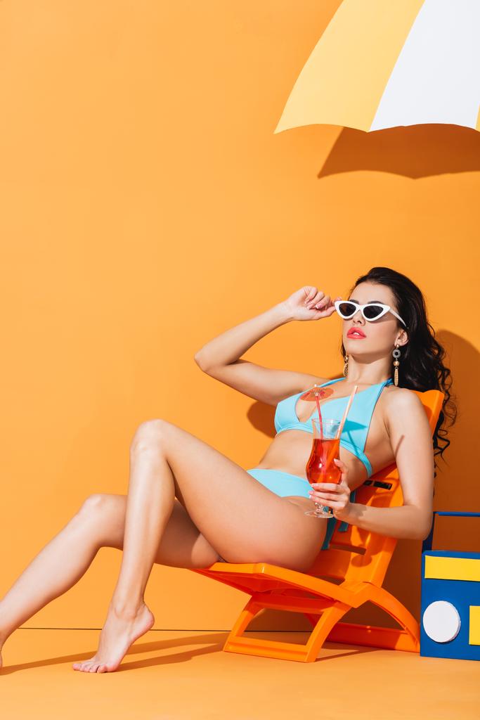 модная женщина в солнечных очках и купальнике сидит на шезлонге возле бумажного бумбокса и зонтика, держа коктейль на апельсине
 - Фото, изображение