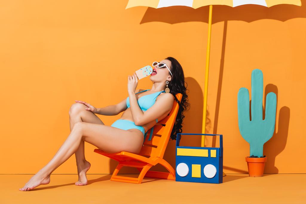 trendi nő napszemüvegben és fürdőruhában ül a fedélzeten szék közelében boombox és esernyő, miközben nyalogatás papír fagylalt narancs - Fotó, kép