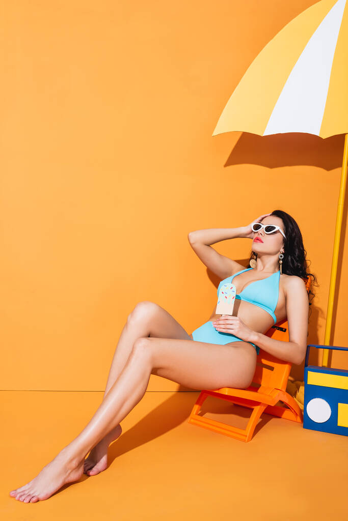 trendy vrouw in zonnebril en badmode zittend op ligstoel in de buurt van papieren boombox en paraplu terwijl ze ijs op oranje houdt - Foto, afbeelding