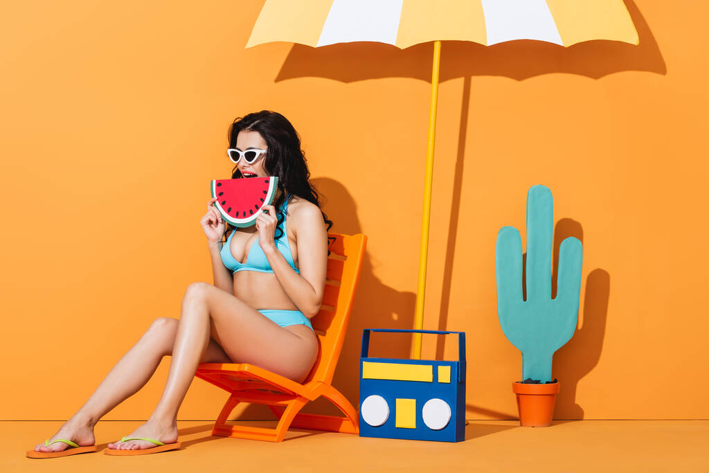 jeune femme en lunettes de soleil et maillot de bain assis sur la chaise longue près de boombox et parapluie tout en tenant pastèque en papier sur orange - Photo, image