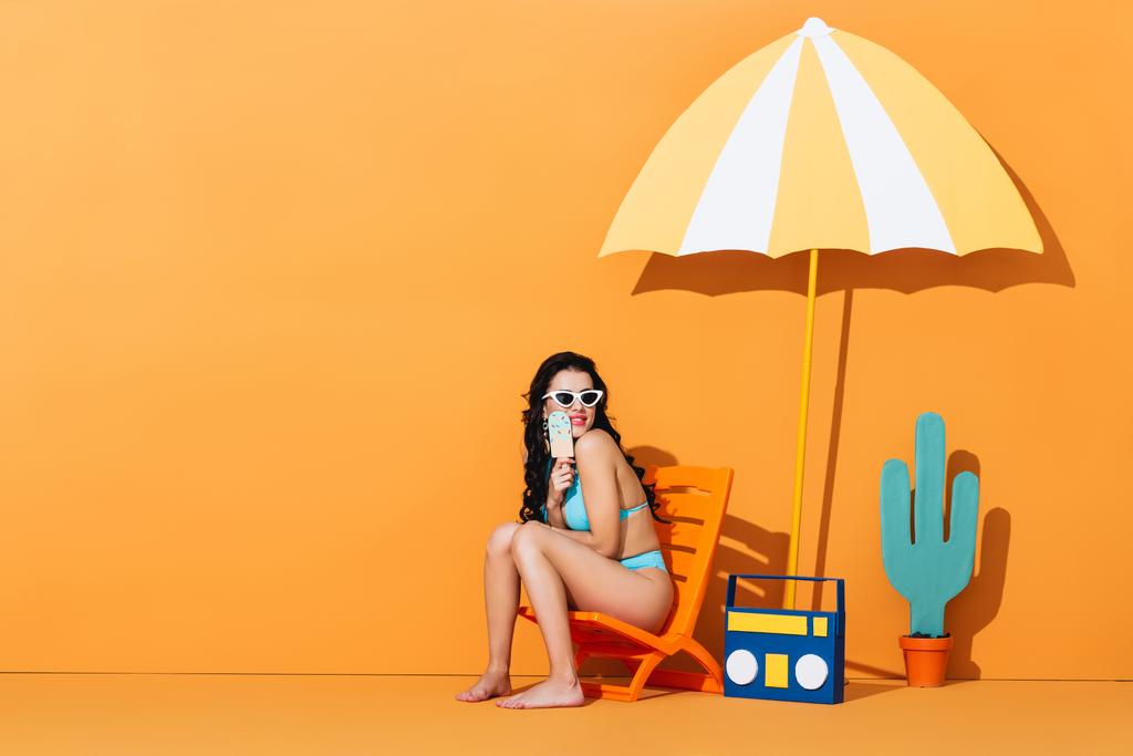happy woman in sunglasses and swimsuit sitting on deck chair near boombox and umbrella while holding paper ice cream on orange - Photo, Image