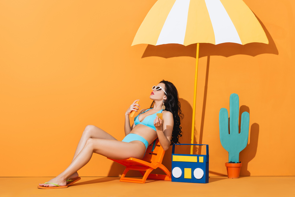 chica con estilo en gafas de sol y traje de baño sentado en la silla de cubierta cerca de la caja de resonancia de papel, cactus y paraguas mientras se aplica protector solar en naranja
 - Foto, Imagen