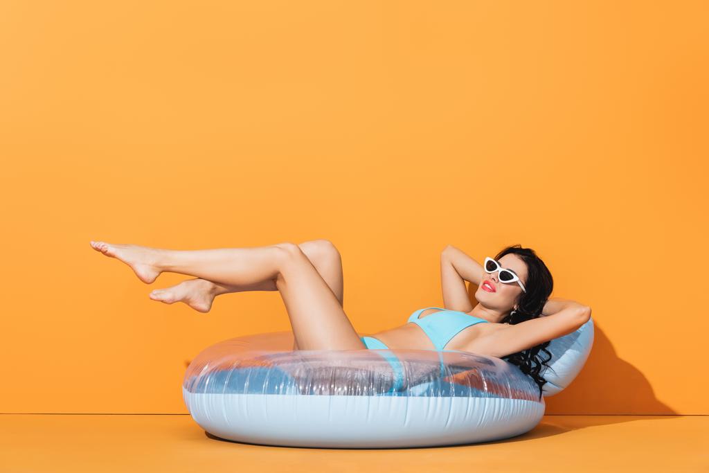 mujer alegre en gafas de sol y trajes de baño acostado en el anillo inflable en naranja
  - Foto, Imagen