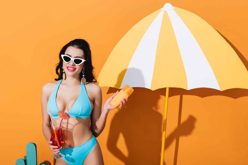glückliche Frau in Badebekleidung und Sonnenbrille mit Sonnencreme und Cocktail in der Nähe eines Papierschirms auf Orange - Foto, Bild