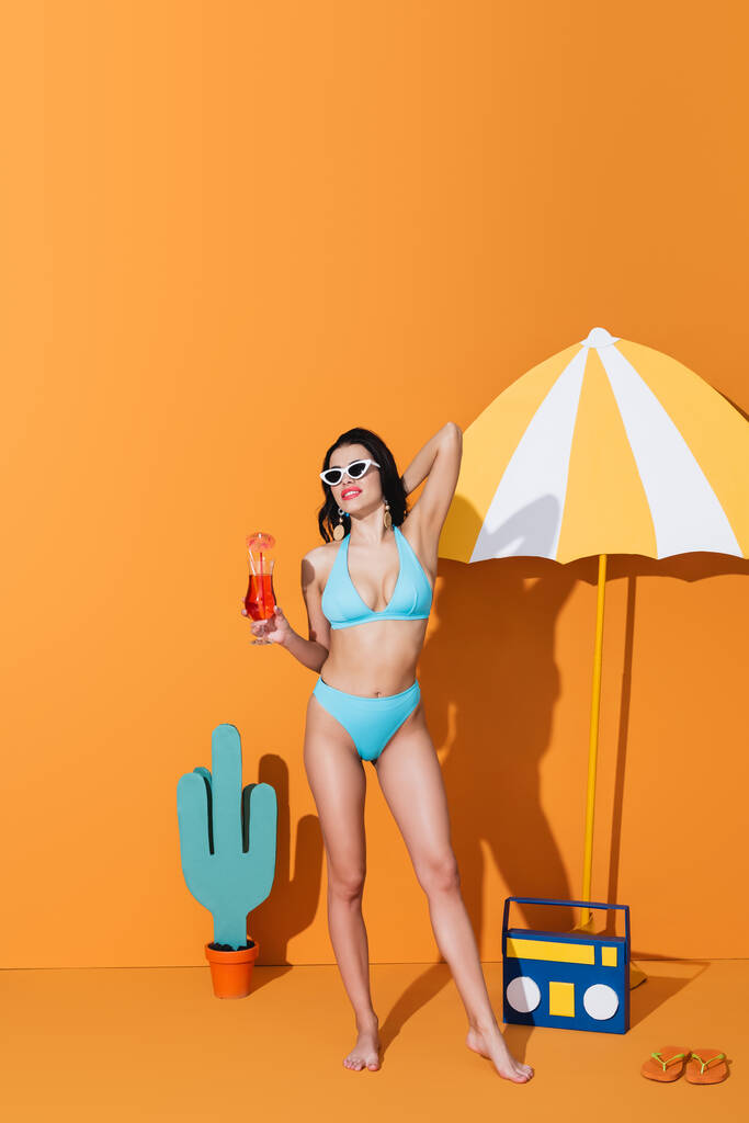 felice donna in costume da bagno e occhiali da sole che tiene cocktail vicino ombrello di carta, boombox e cactus su arancione - Foto, immagini