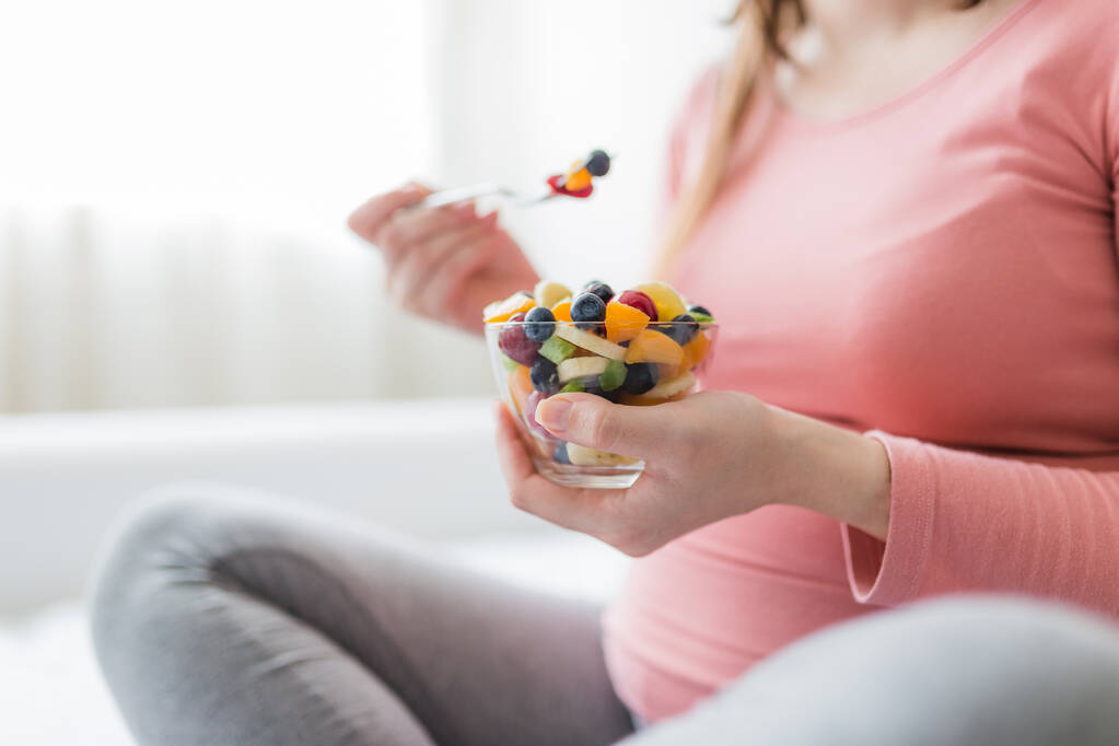 Közelkép egy terhes nőről, aki az ágyon ül és gyümölcssalátát eszik. Egészséges étkezés koncepciója. - Fotó, kép