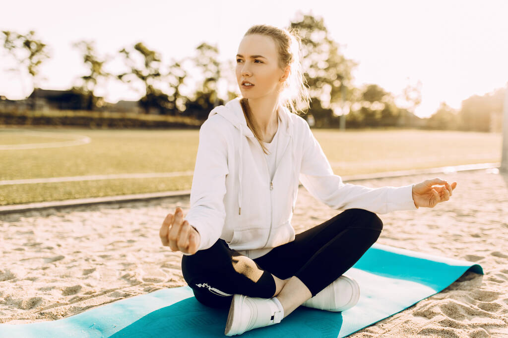 mujer deportiva practica yoga y meditación en la posición de Loto mientras está sentada en una alfombra de fitness en la playa por la mañana
 - Foto, Imagen