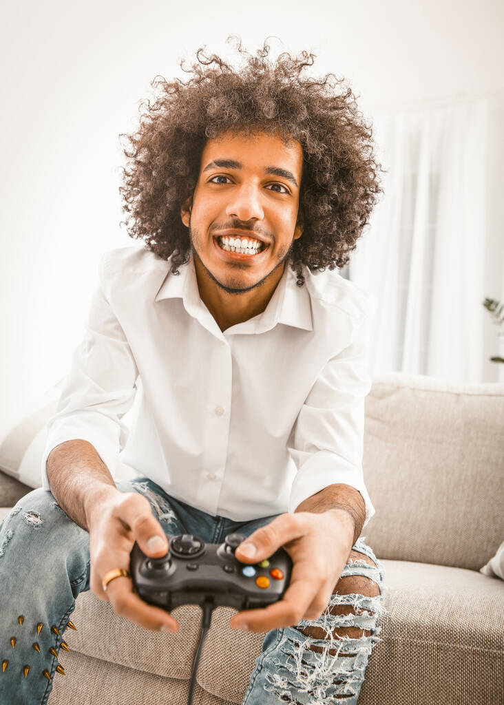 Młody człowiek gra w grę komputerową sam w domu. Shuggy gracz uśmiecha się trzymając joysticka. Koncepcja samoizolacji. Holenderski kąt widzenia. Koncepcja emocji. Przyciemniany obraz - Zdjęcie, obraz
