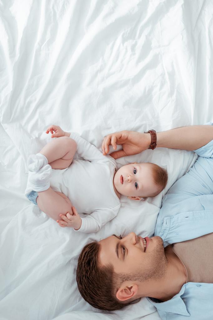 top view of happy young man touching adorable baby boy while lying near him in bed - Photo, Image