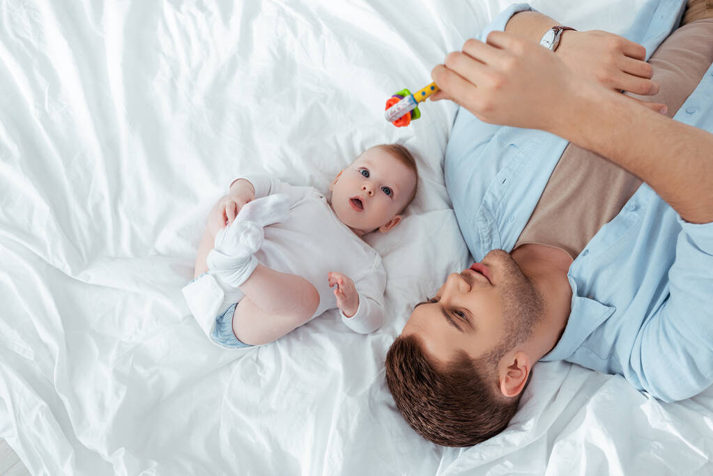κορυφαία άποψη του νεαρού πατέρα παίζει με κουδουνίστρα μωρό πάνω χαριτωμένο μικρό γιο που βρίσκεται στο κρεβάτι - Φωτογραφία, εικόνα