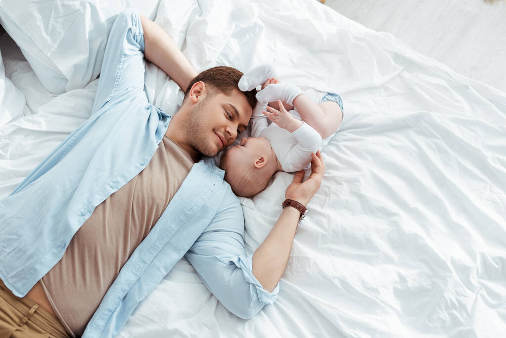 κορυφαία άποψη του ευτυχισμένος πατέρας ξαπλωμένος στο κρεβάτι πρόσωπο με πρόσωπο με χαριτωμένο αγοράκι - Φωτογραφία, εικόνα