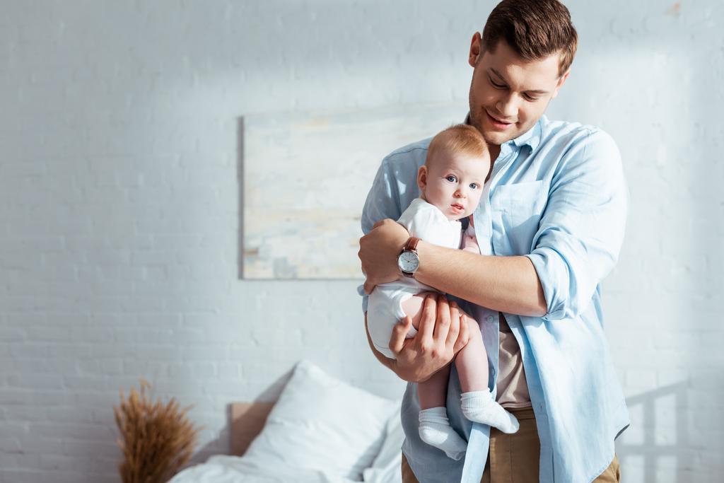 glücklicher junger Vater hält und umarmt entzückenden kleinen Sohn im Schlafzimmer - Foto, Bild