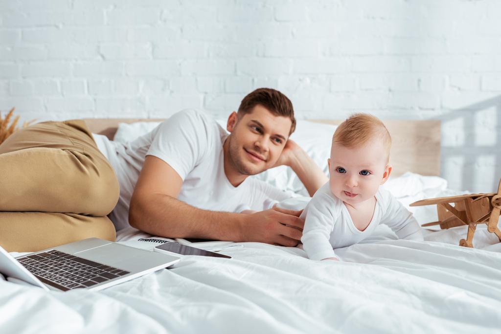 可愛い息子やノートパソコンやおもちゃの飛行機のそばのベッドに横になった幸せな男 - 写真・画像