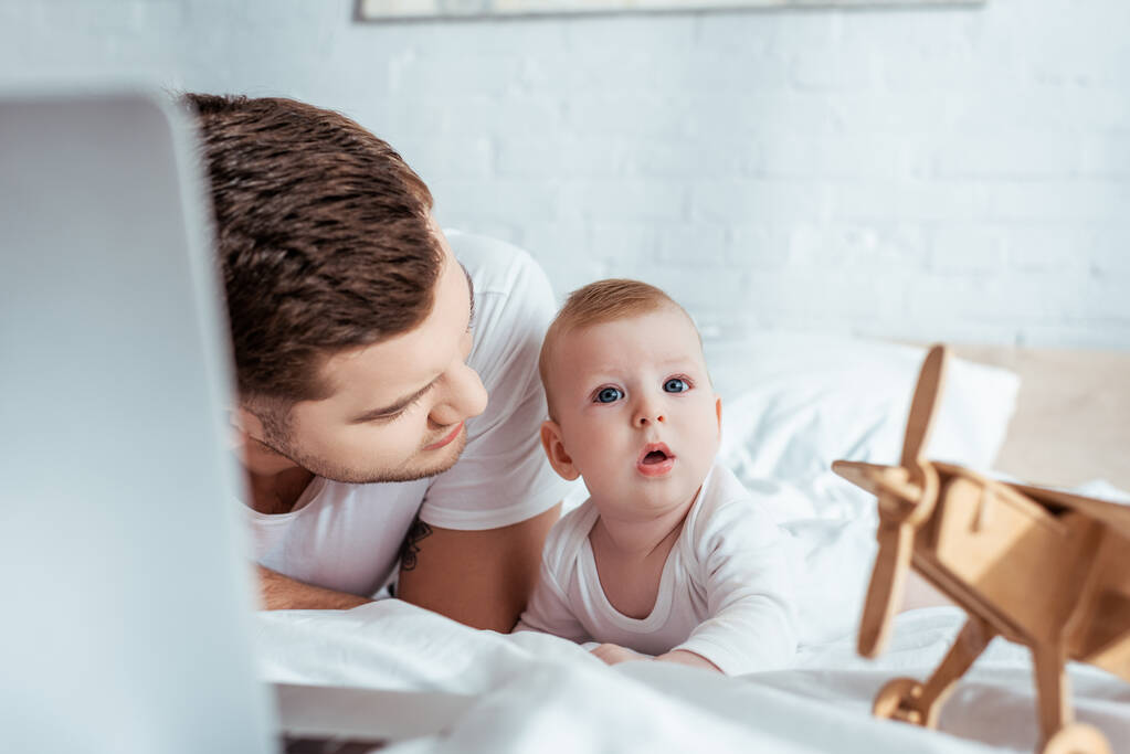 enfoque selectivo de padre joven mirando sorprendido bebé niño mientras está acostado en la cama cerca de la computadora portátil y avión de juguete
 - Foto, imagen