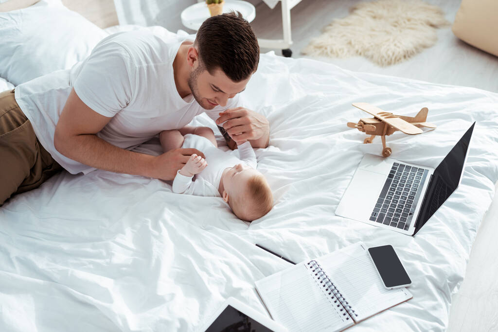 Junger Vater spielt mit Baby in der Nähe von Gadgets, Notizbuch und Spielzeugflugzeug im Bett - Foto, Bild