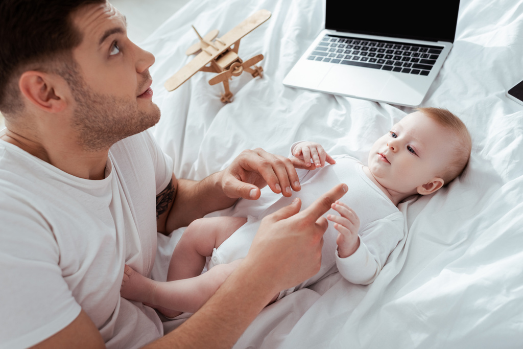 молодой отец смотрит вверх и трогает малыша рядом с ноутбуком и игрушечным самолетом
 - Фото, изображение