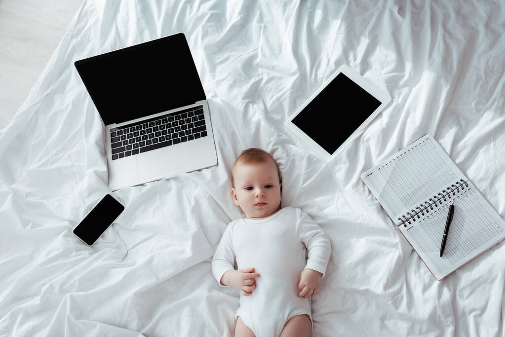 vrchní pohled na roztomilé dítě ležící na posteli v blízkosti gadgets a notebook s perem - Fotografie, Obrázek