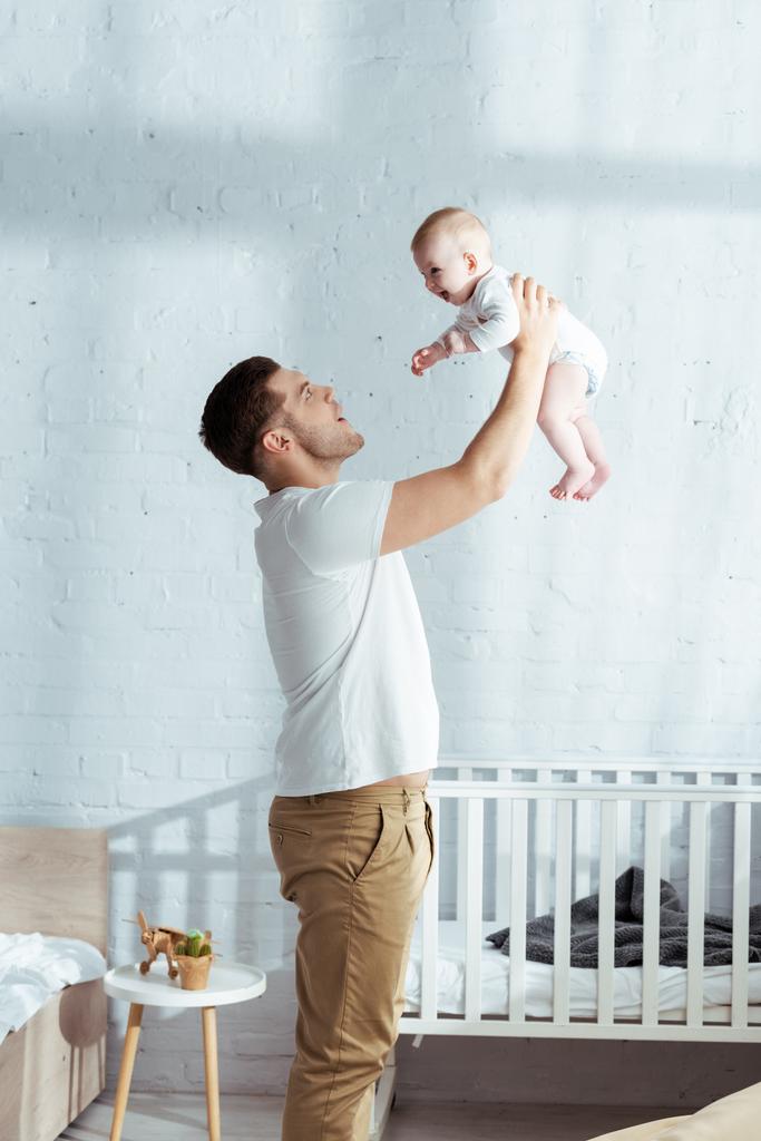 happy father holding adorable baby boy in outstretched hands near baby cot - Photo, Image