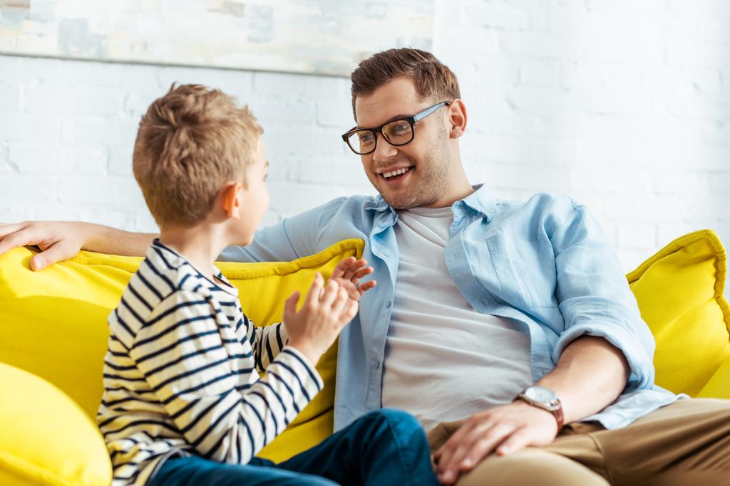 smiling man talking to adorable son while sitting on sofa at home - Photo, Image