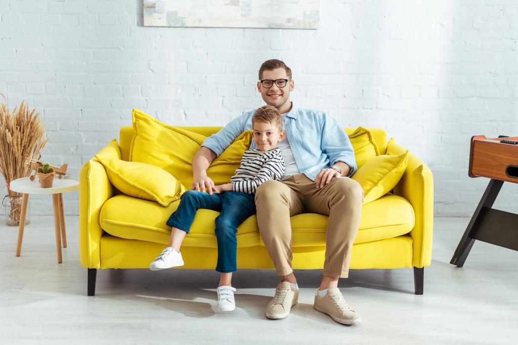szczęśliwy ojciec i syn patrząc w aparat podczas siedzenia na żółtej kanapie - Zdjęcie, obraz