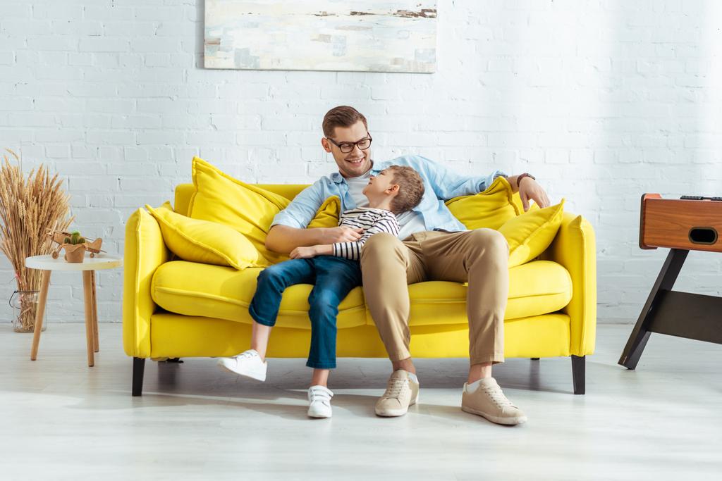 Χαρούμενος πατέρας και γιος μιλάνε ενώ κάθονται στον κίτρινο καναπέ - Φωτογραφία, εικόνα