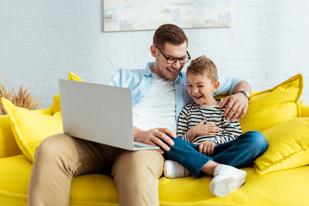 opgewonden vader en zoon lachen terwijl zitten op de gele bank met laptop - Foto, afbeelding