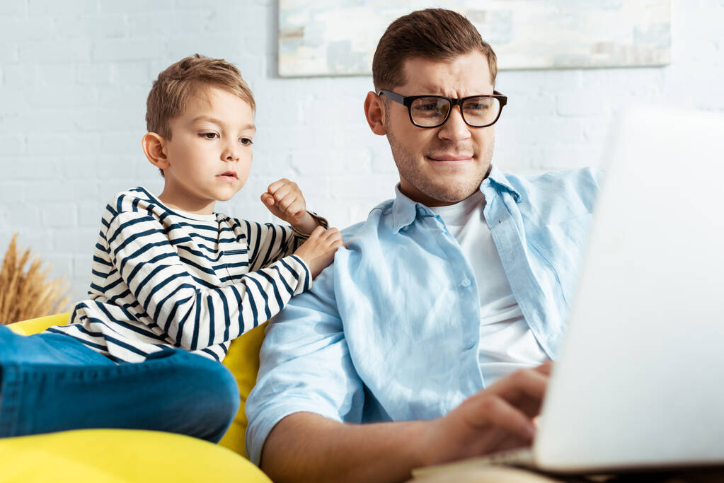niño lindo sentado cerca de padre atento usando el ordenador portátil
 - Foto, imagen