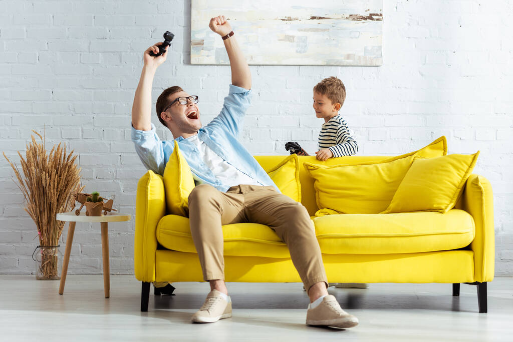 KYIV, UKRAINE - JUNE 9, 2020: excited man showing winner gesture while playing video game with son - Photo, Image