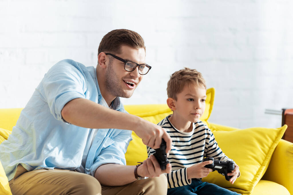 KYIV, UCRANIA - 9 de junio de 2020: padre emocionado e hijo atento jugando videojuegos con joysticks
 - Foto, imagen