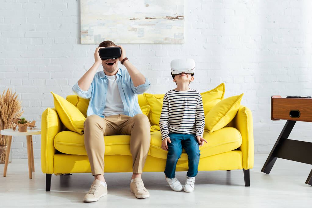 excité père et fils en utilisant des casques vr tout en étant assis sur le canapé jaune - Photo, image