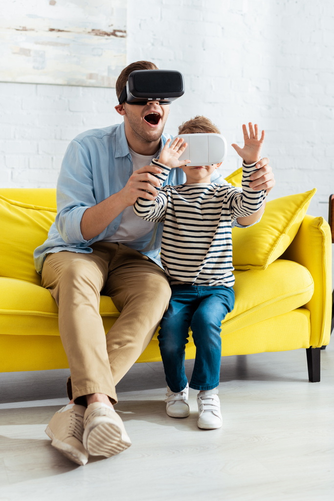 aufgeregter Mann hält Hände seines Sohnes, während er VR-Headsets zusammen benutzt - Foto, Bild