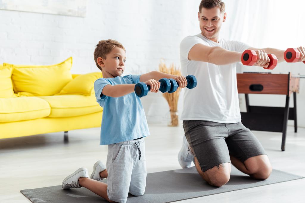 père souriant avec fils faisant de l'exercice avec des haltères tout en se tenant sur les genoux sur tapis de fitness - Photo, image