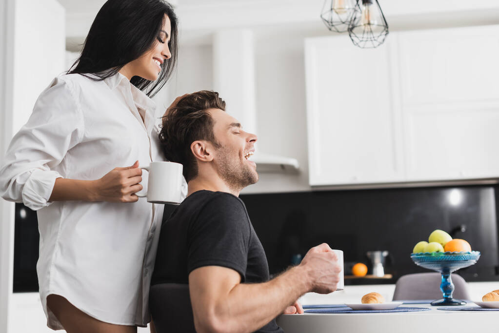 Boční pohled na sexy žena v košili drží šálek kávy v blízkosti veselý přítel v kuchyni  - Fotografie, Obrázek