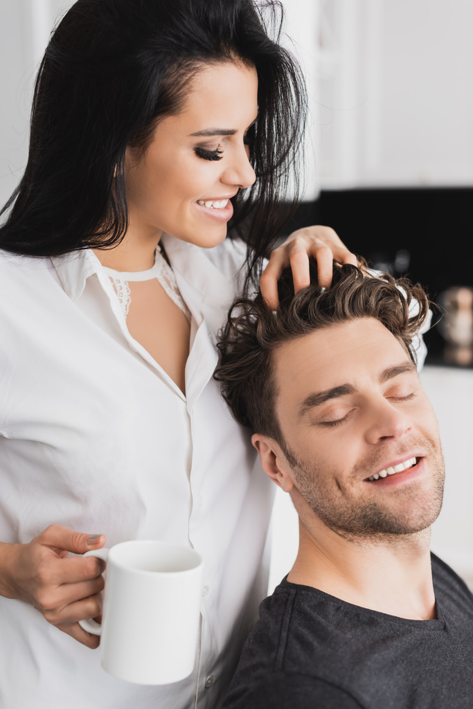 Uśmiechnięta kobieta w koszuli trzyma filiżankę kawy i dotyka włosów przystojnego chłopaka  - Zdjęcie, obraz