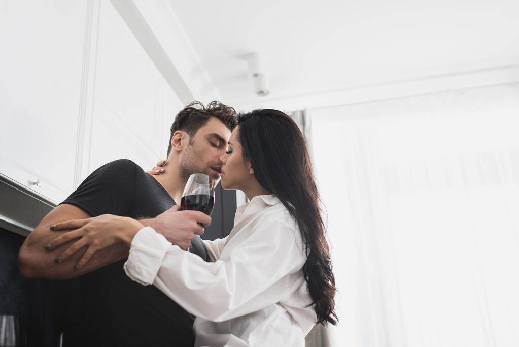 Donna in camicia baciare bell'uomo con bicchiere di vino in cucina  - Foto, immagini