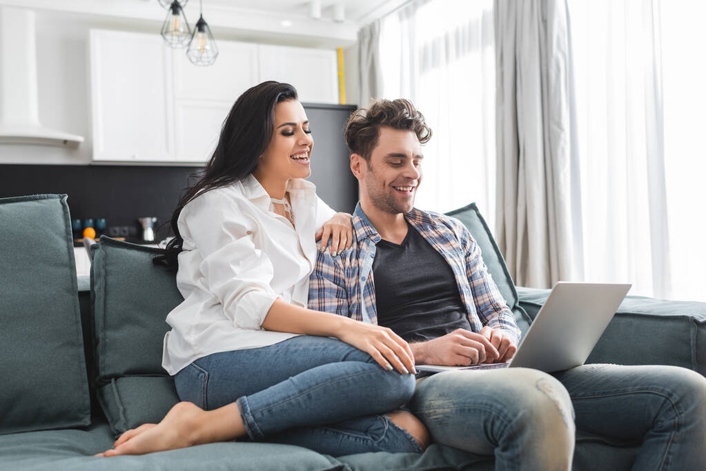 Sorrindo mulher sentada perto bonito namorado usando laptop no sofá em casa
  - Foto, Imagem